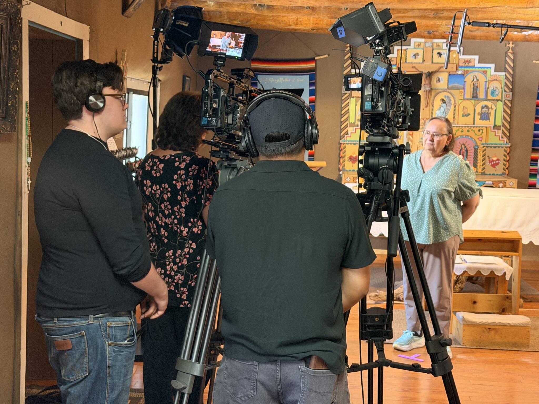 Video production crew filming an interview: multiple cameras on tripods facing a woman in a teal blouse indoors.