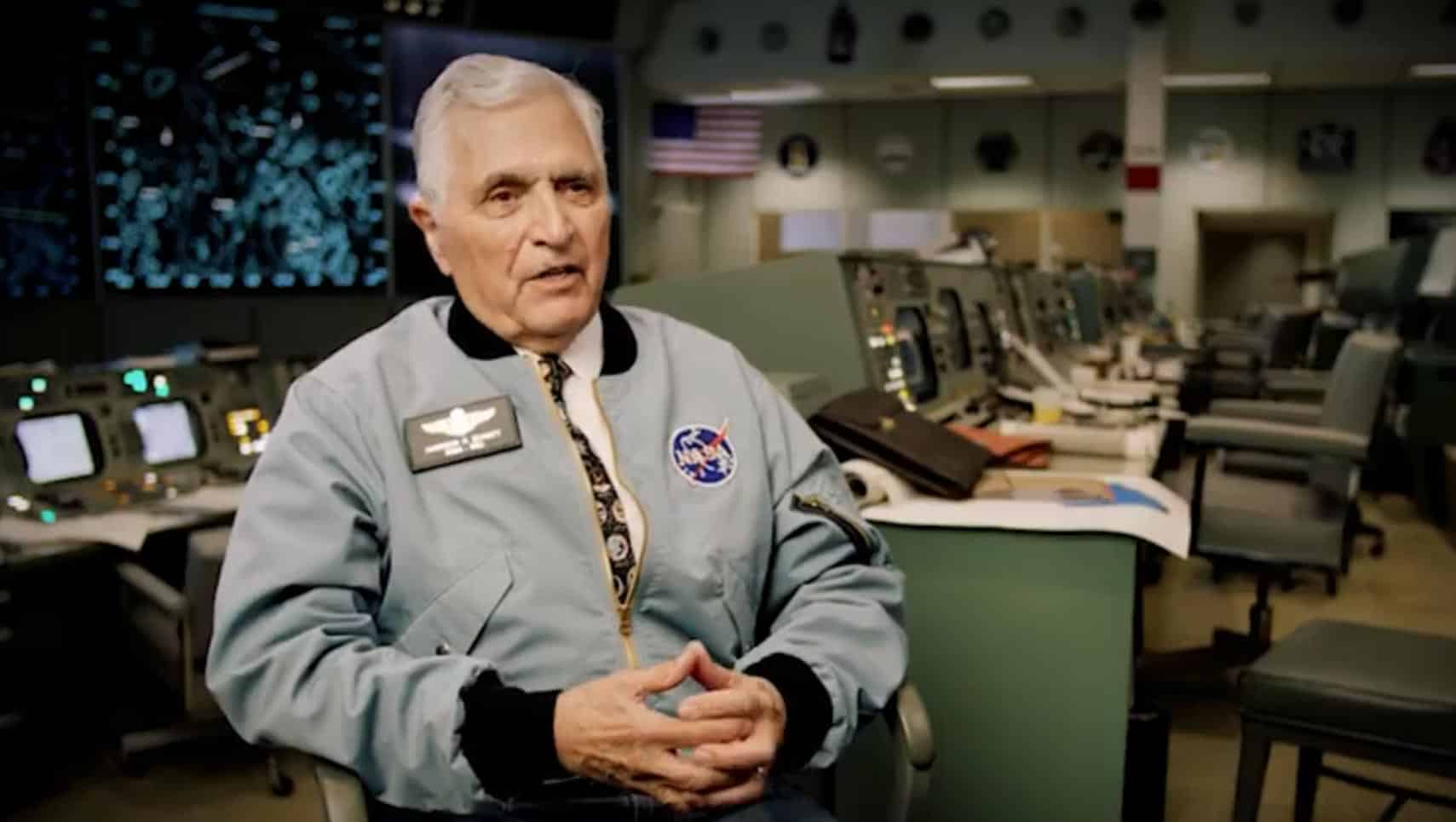 Older man in a light blue NASA-style jacket with a badge, seated in a spacecraft control room with consoles and screens behind him.