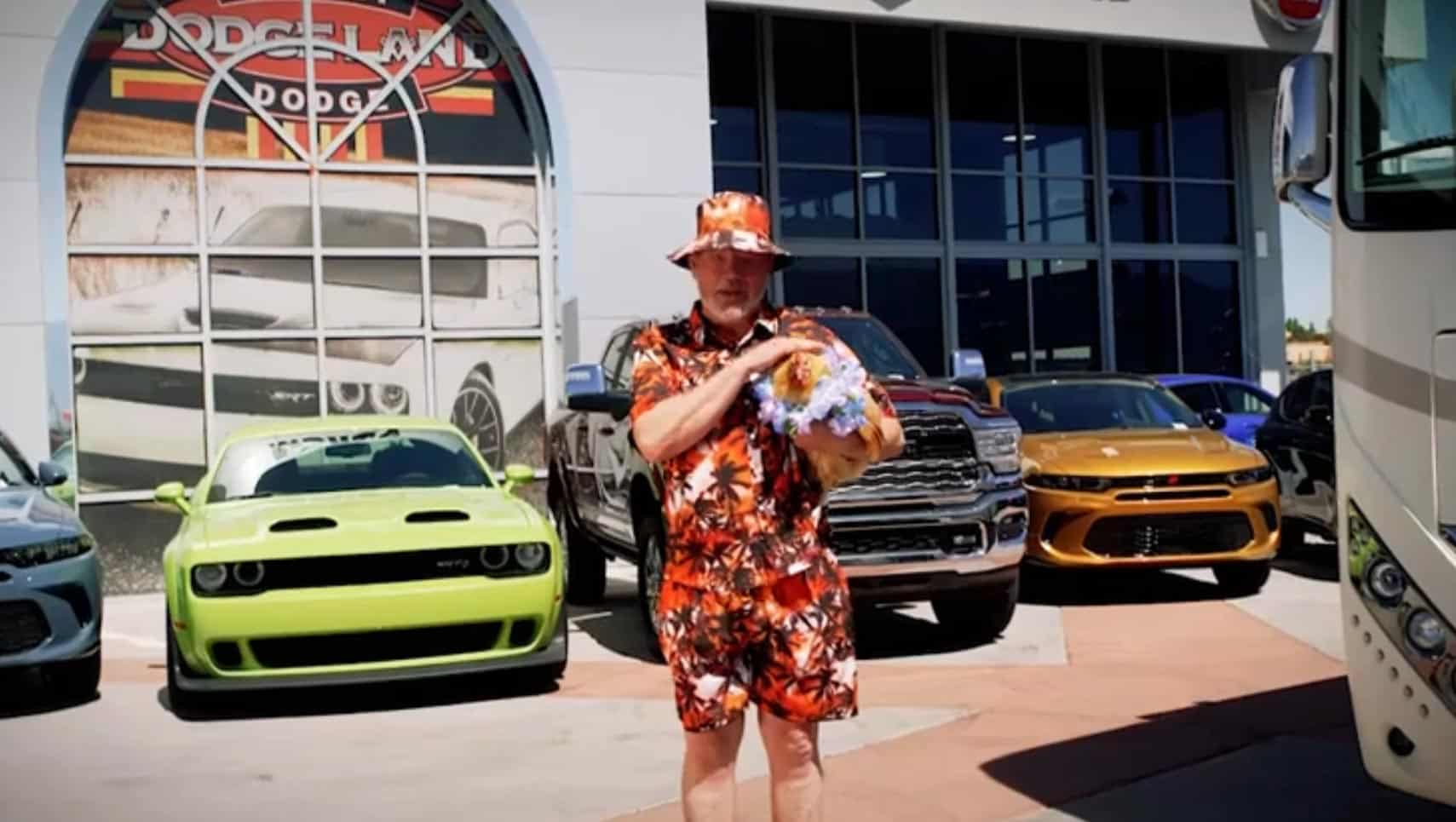 Man in a bright floral outfit holding a bouquet, standing in front of colorful cars at a dealership.