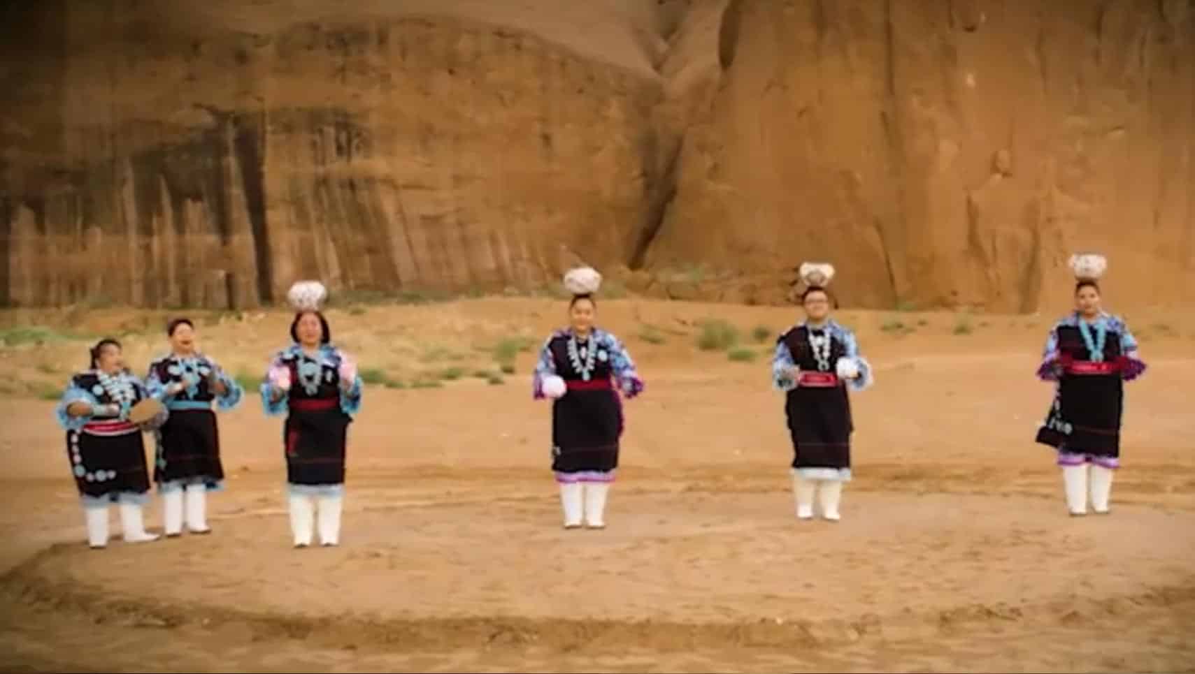 A line of women in colorful traditional outfits dancing on a sandy shore with a rocky cliff in the background.