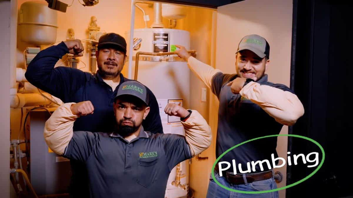 Three male plumbers in uniform flexing their biceps in a boiler room with a water heater behind them, smiling.