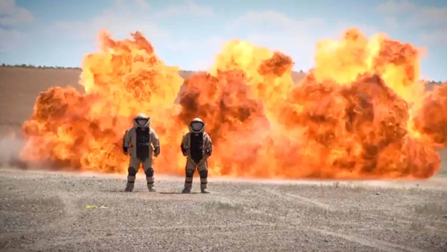 Two astronauts in space suits stand facing a massive orange explosion in a barren desert landscape.