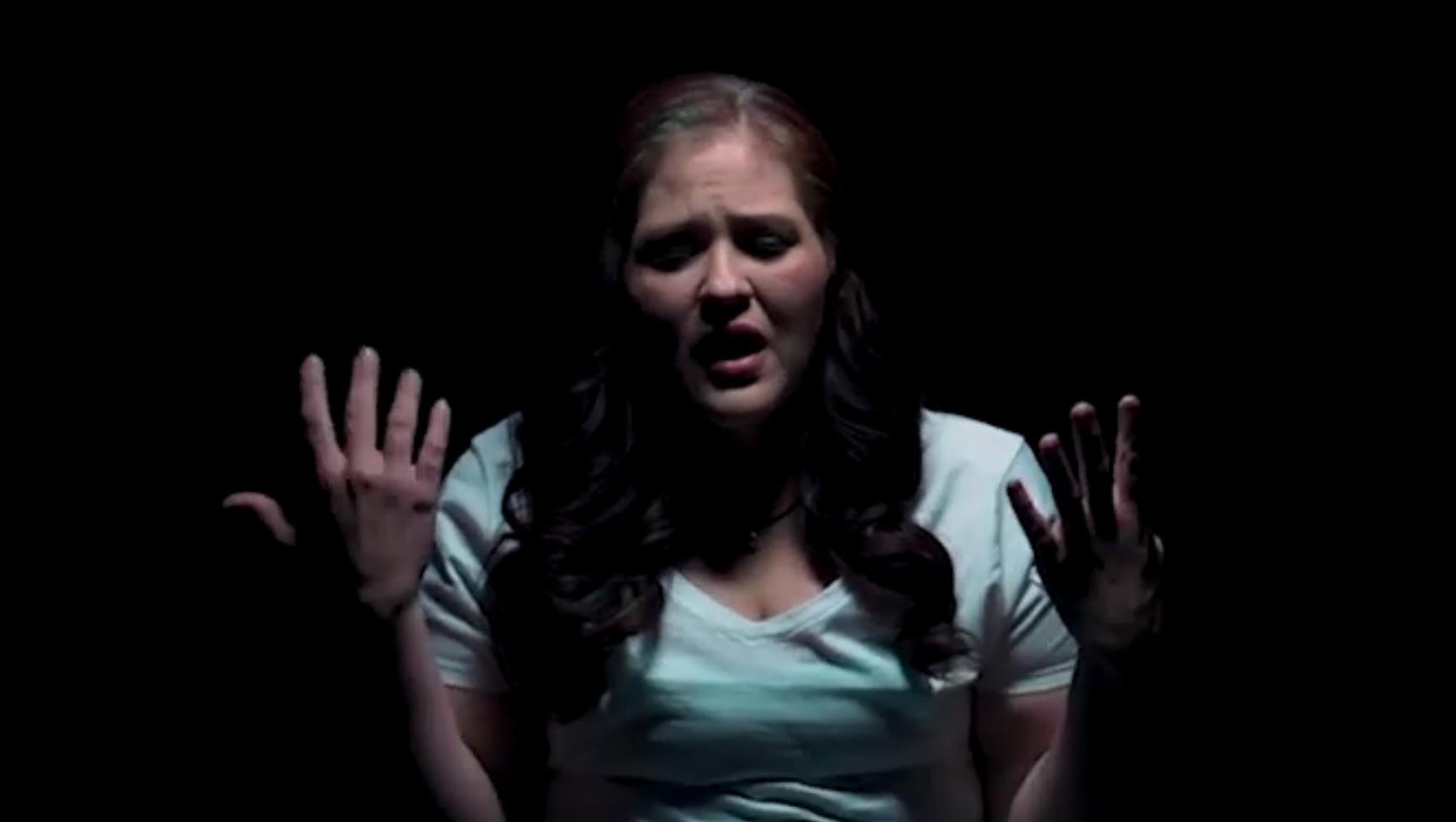 Woman singing with hands raised against a dark background, dramatic spotlight on her face and upper body.