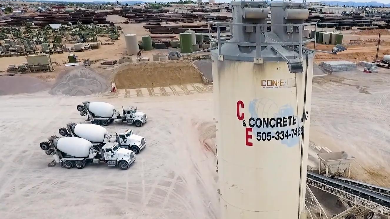 Aerial view of a cement plant with three concrete mixer trucks parked near a tall silo and surrounding storage yards.